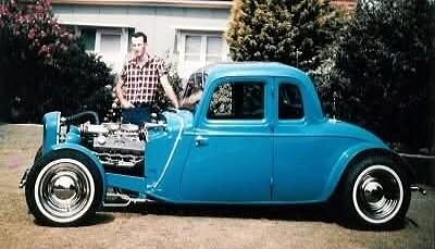 A man stands proudly next to a custom blue hot rod car with an exposed engine and classic white-wall tires.