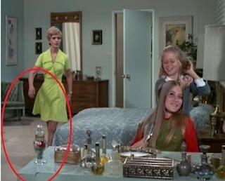 A woman in a green dress stands in the background, while two younger women sit at a vanity table in the foreground. A decorative bottle is highlighted by a red circle on the table.