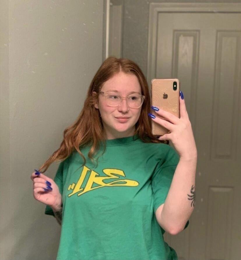 A woman taking a mirror selfie. She has red hair, wears glasses, and a green t-shirt with a yellow '311' logo. She has blue nail polish and a nose piercing.
