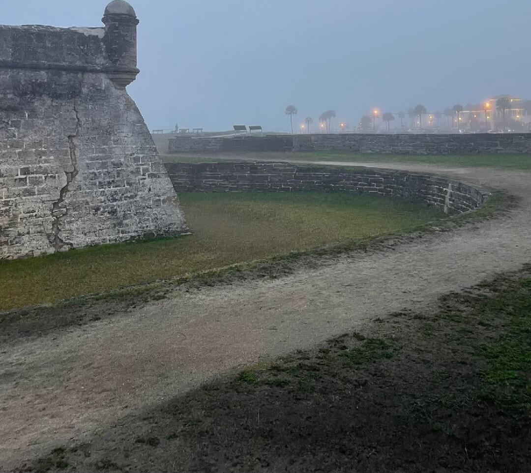 A dirt path along the inside of a stone fortress wall, with a view of the waterfront and distant lights at dusk.