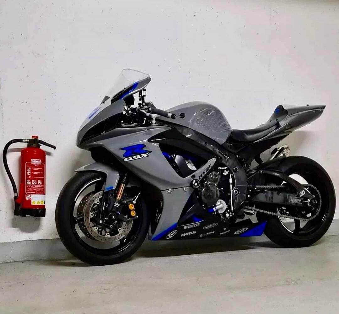 A grey and blue Suzuki GSX-R motorcycle is parked next to a red fire extinguisher. The motorcycle features various sponsor decals including ECSTAR, MOTUL, PIRELLI, ARROW, brembo, and OHLINS.