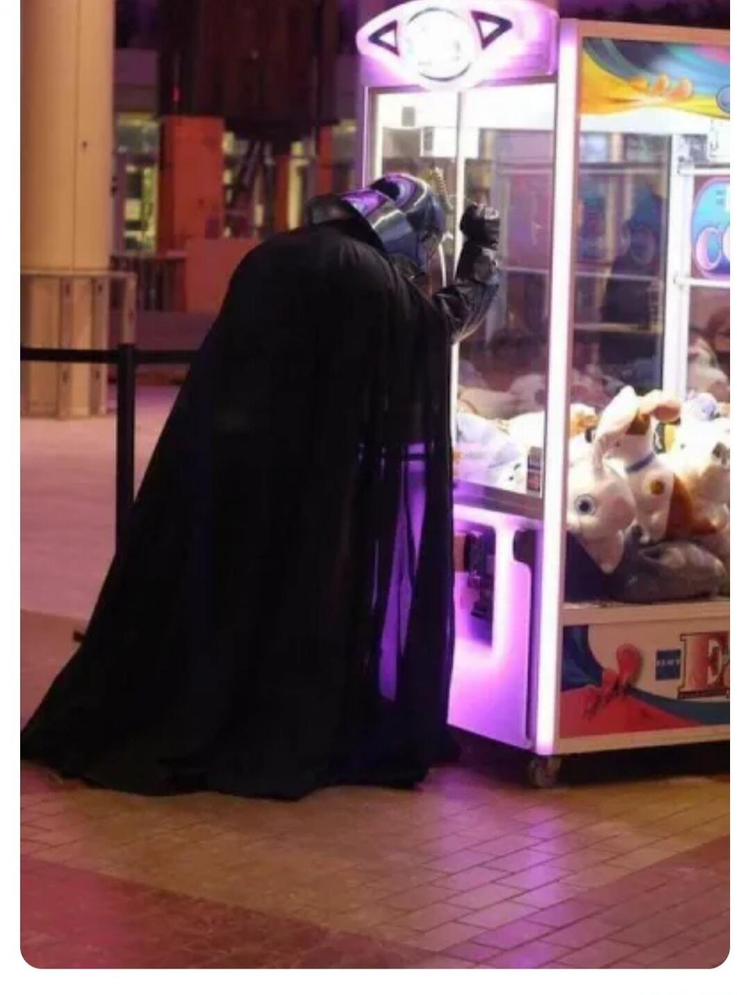 A person dressed as Darth Vader is playing a claw machine filled with plush toys in what appears to be an arcade or mall.