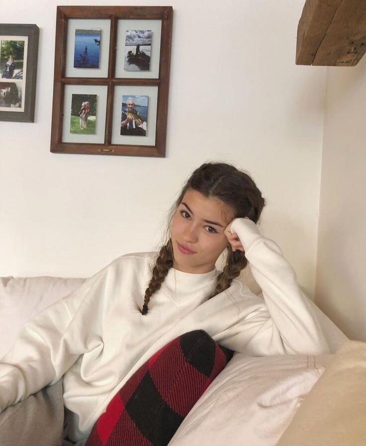 A woman with braids wearing a white sweater sitting on a couch; framed photos on the wall behind her.