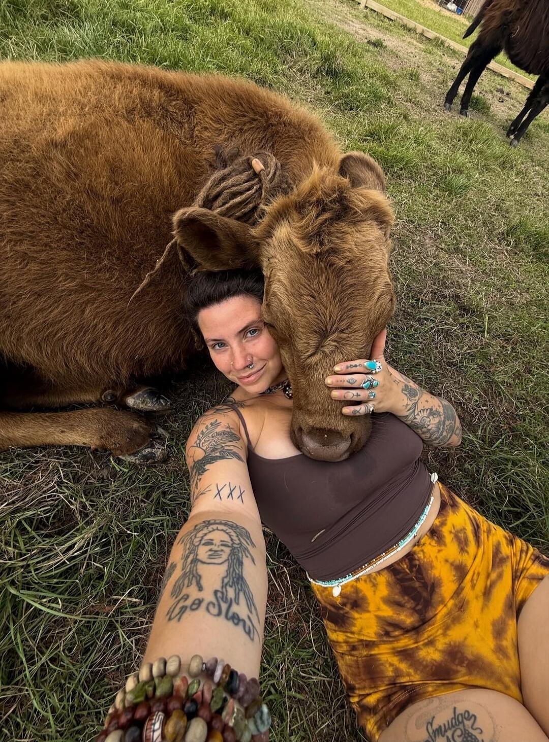 A woman with tattoos, including 'Go Slow', 'XXX', and 'Smudge', is lying on the grass, cuddling a brown calf. Another cow is visible in the background.
