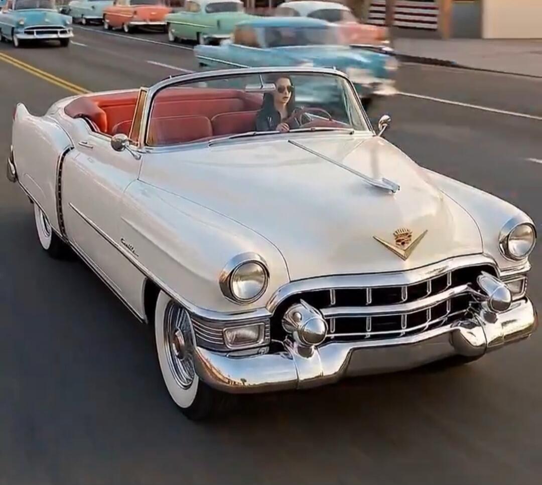 A woman in sunglasses drives a white vintage Cadillac convertible with a red interior down a road. Several other classic cars are visible in the background, suggesting a parade or car show.