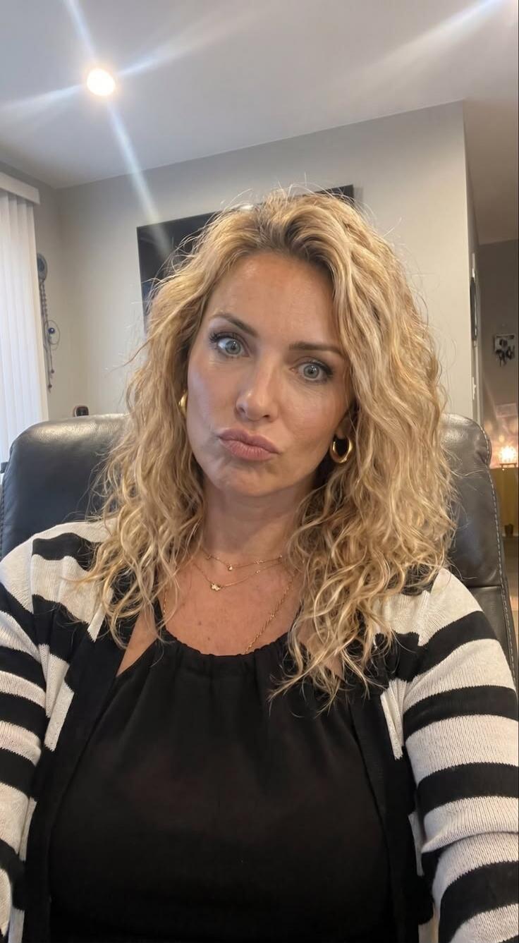 A woman with blonde curly hair and blue eyes making a pouty face, wearing a black top and a striped cardigan.