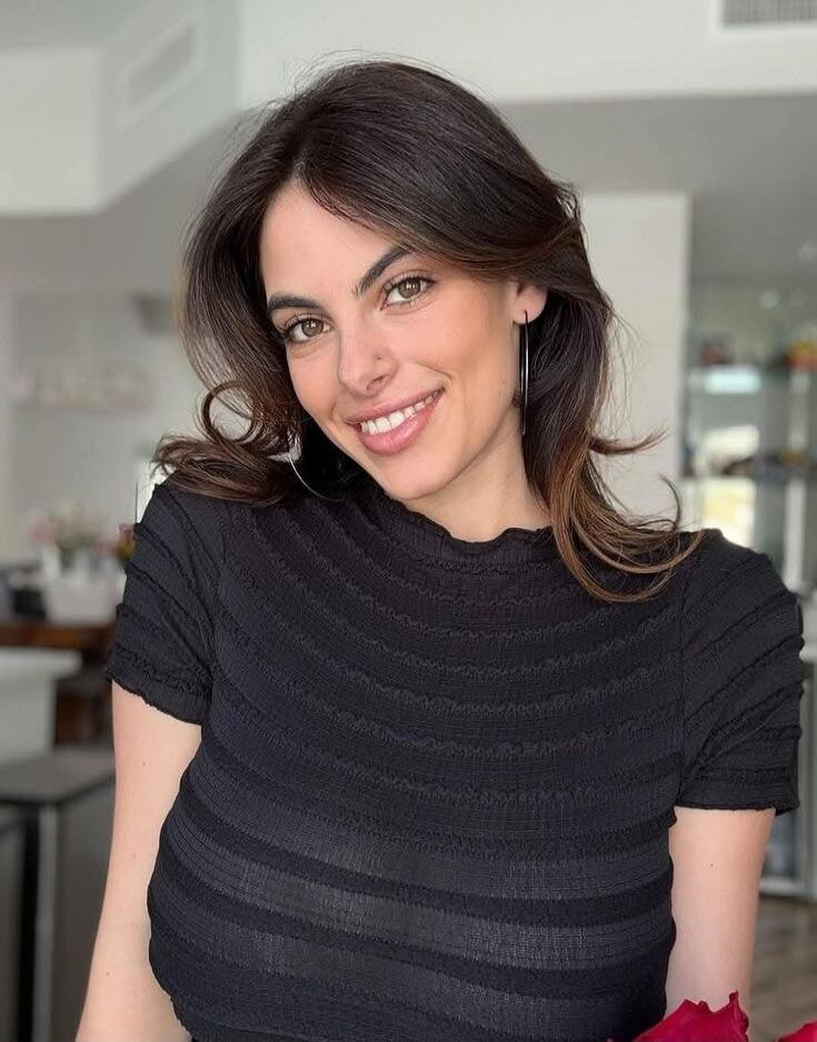 A woman with shoulder-length brown hair, wearing a black striped top, smiling indoors.