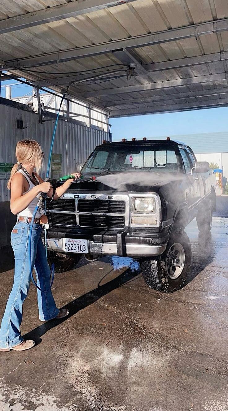 A woman is washing a black Dodge pickup truck at a car wash. The truck has a California license plate with '52237D3'. A 'MENU' sign is visible in the background.