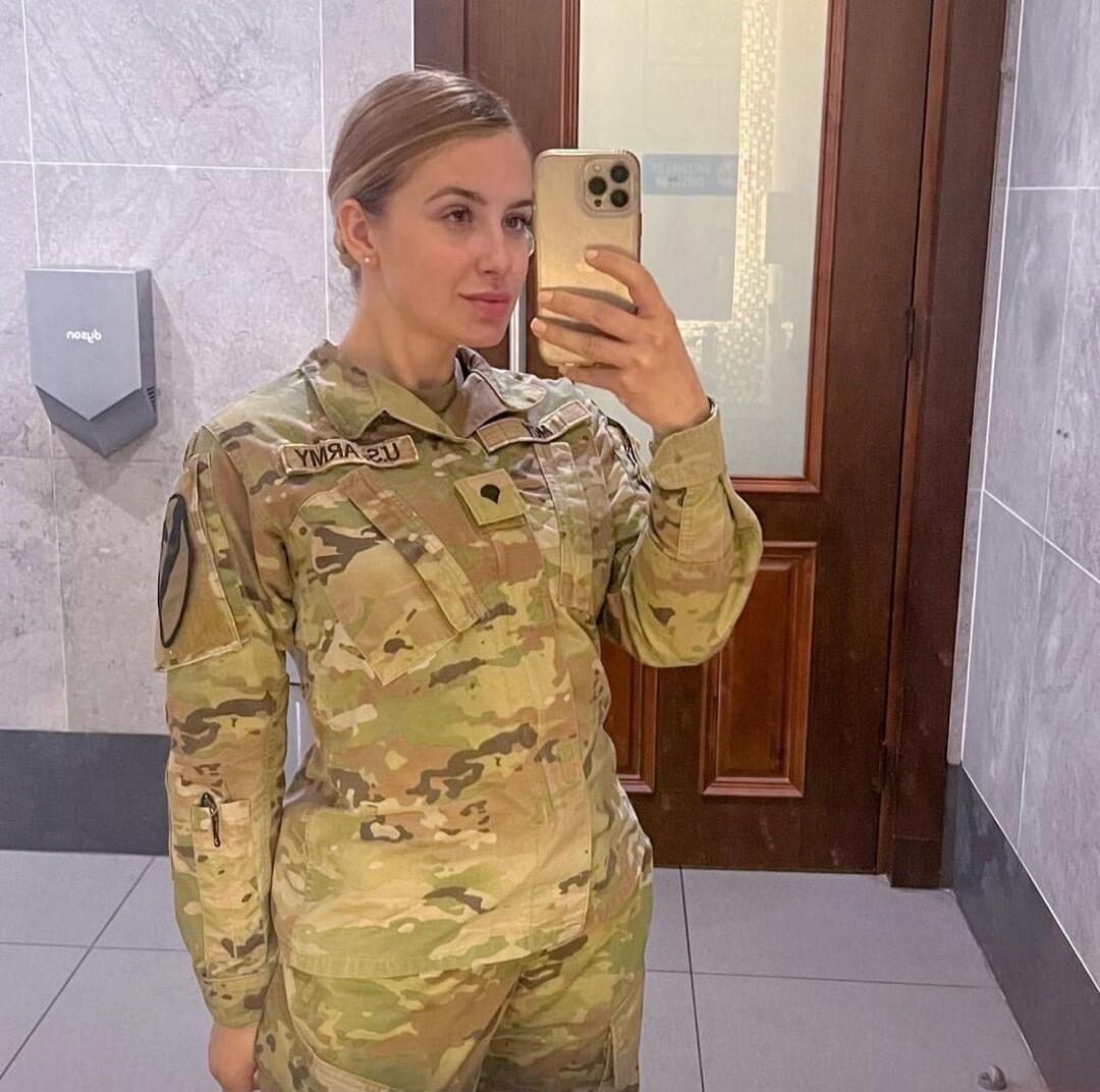 A woman in camouflage military uniform taking a mirror selfie in a bathroom.