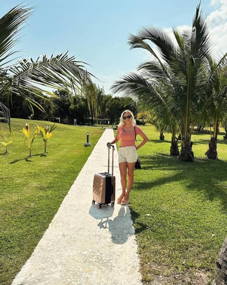 A woman standing on a white stone path in a tropical garden with palm trees. She is wearing a pink top, white shorts, sandals, and sunglasses, and has a rolling suitcase beside her.