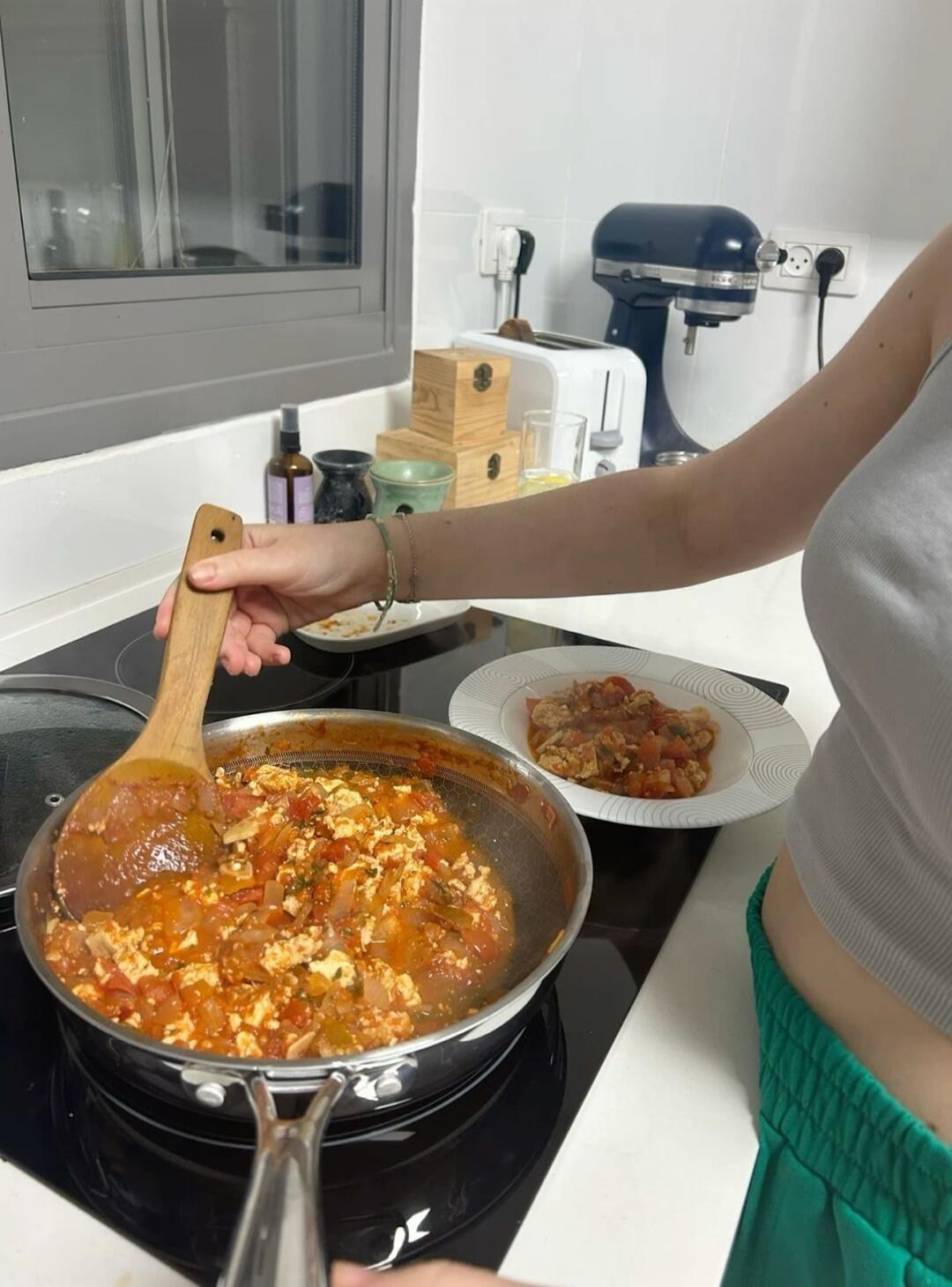 A person stirs a tomato-based sauce in a pan on a stove, using a wooden spatula. There is a plate with more food in the background and various kitchen items on the counter.