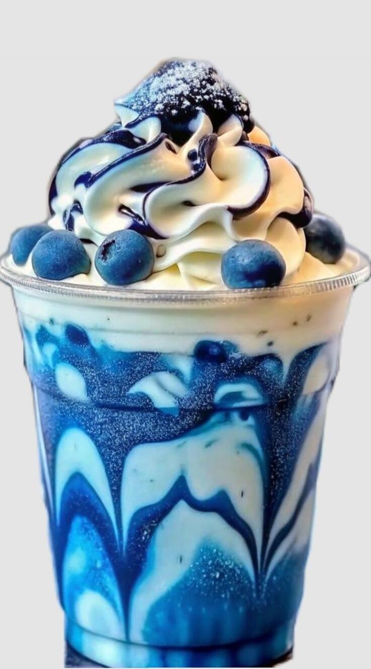 A vibrant blue and white layered blueberry milkshake or smoothie in a clear cup, topped with whipped cream, blueberry syrup, and fresh blueberries.