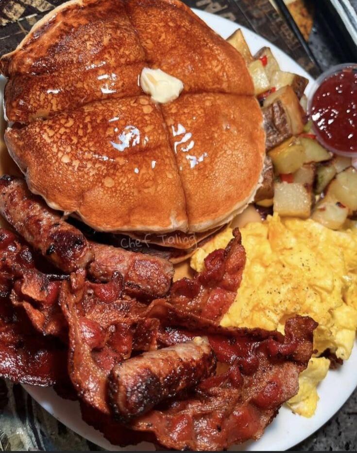 A hearty breakfast plate featuring pancakes with a dab of butter, crispy bacon, scrambled eggs, home fries, and a side of ketchup.