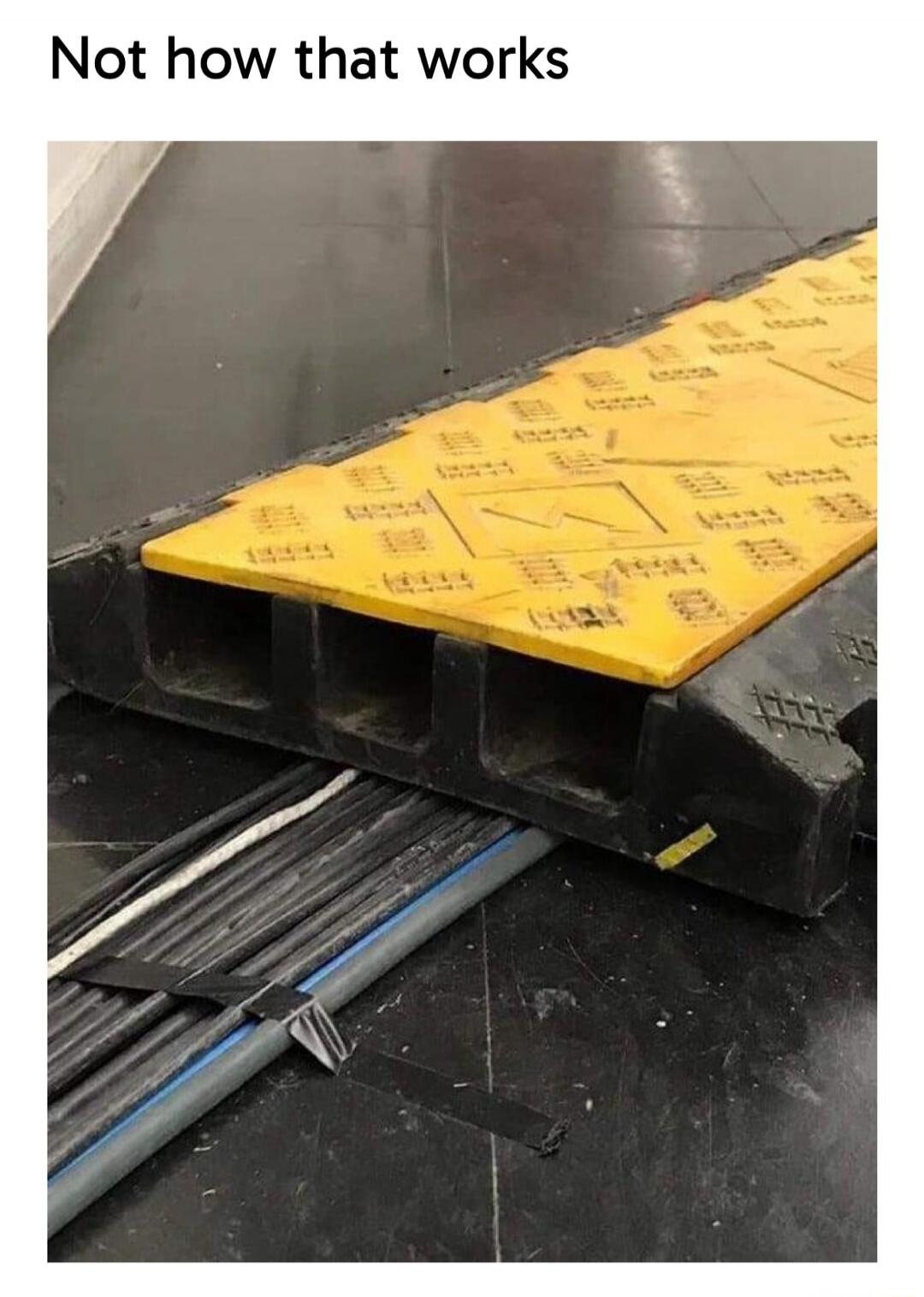 Not how that works. A yellow and black cable protector is placed on the floor, but the electrical cables it is supposed to protect are laid next to it and partially underneath, not properly routed through its channels. This shows a clear misuse of the equipment.