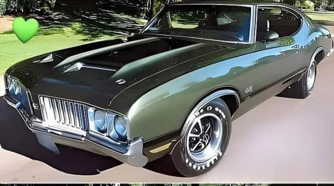 A classic dark green Oldsmobile 442 muscle car is parked outdoors, with a green heart emoji in the top left corner.
