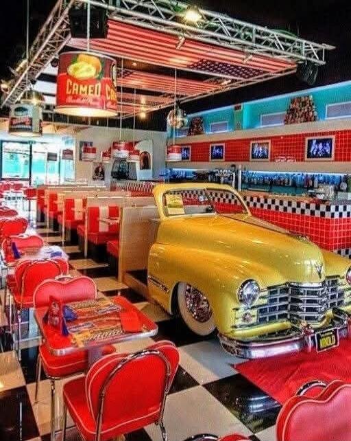A retro 1950s diner interior with a bright yellow vintage car as the centerpiece, red booths, red chairs, checkered black-and-white floor, neon signs, and a long counter.