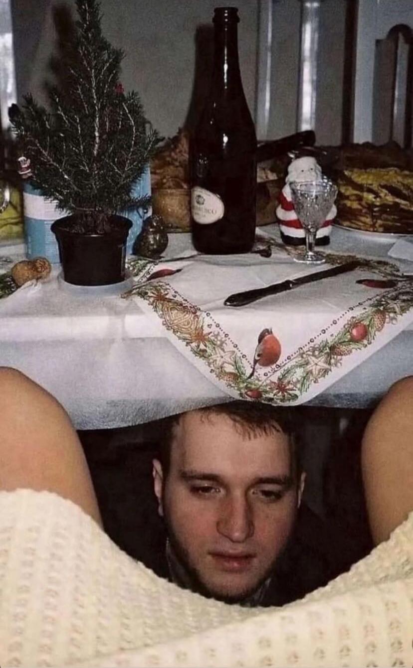 A man is peeking from under a table at a festive setting. On the table there is a small potted Christmas tree, a bottle, a Santa figurine, a knife on a decorative tablecloth, and various party items.