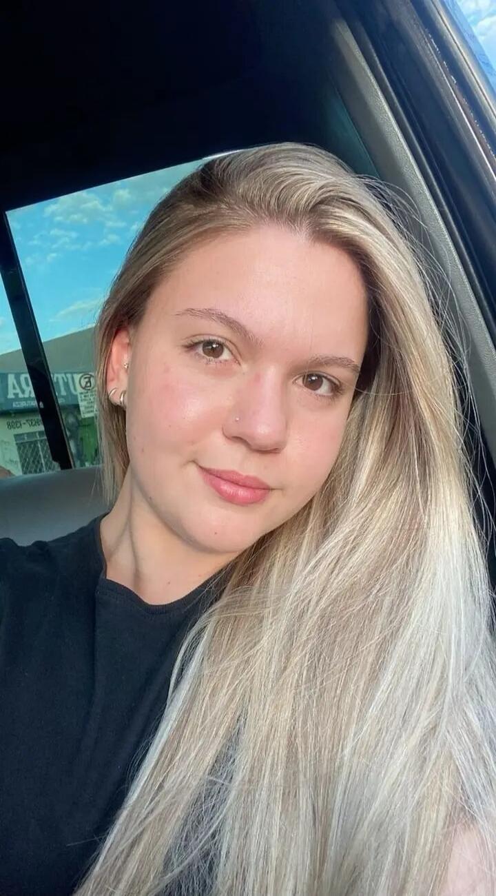 A young woman with long blonde hair and a slight smile is looking at the camera. She is wearing a black t-shirt and has a nose piercing and ear piercings. The background shows a reflection of the sky and some mirrored text through a car window.