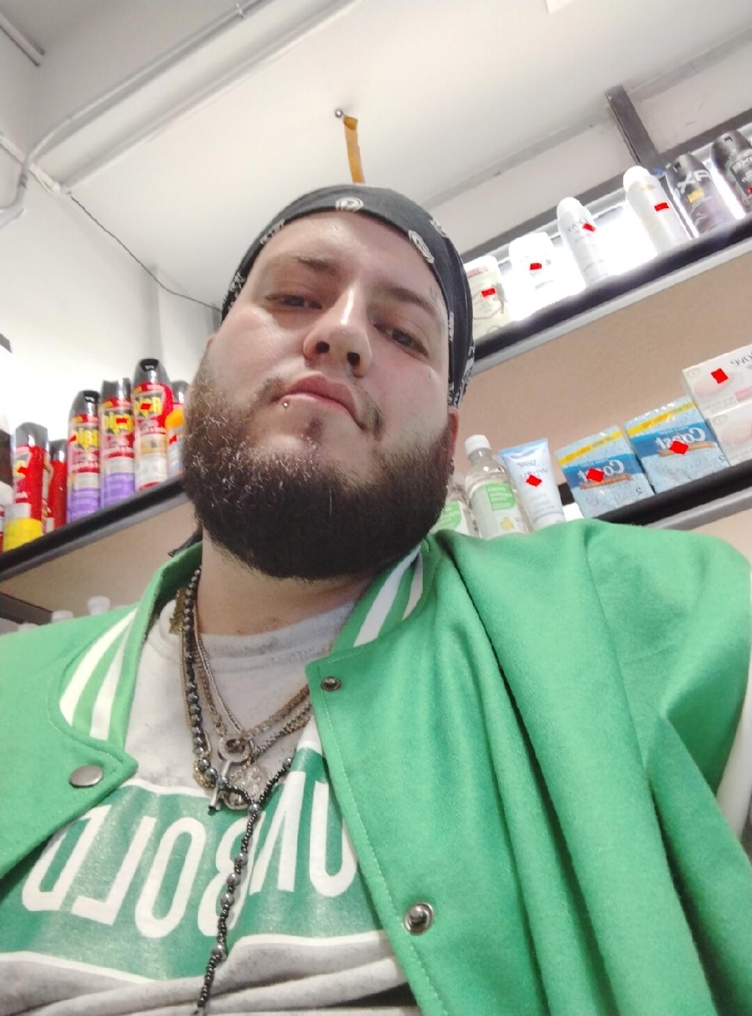 A man with a beard and a lip piercing, wearing a bandana, a green jacket, and multiple necklaces, takes a selfie in what appears to be a store aisle with shelves of products. The text 'BORO' is partially visible on his t-shirt.