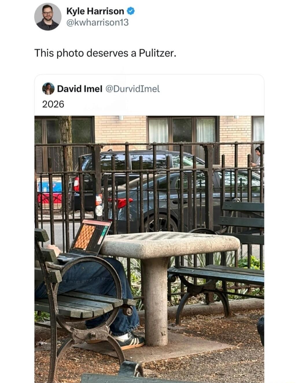 Kyle Harrison @kwharrison13 This photo deserves a Pulitzer. David Imel @DurvidImel 2026. A person is sitting on a park bench, looking at a laptop with a chess-like pattern on the screen, next to an empty stone chess table.