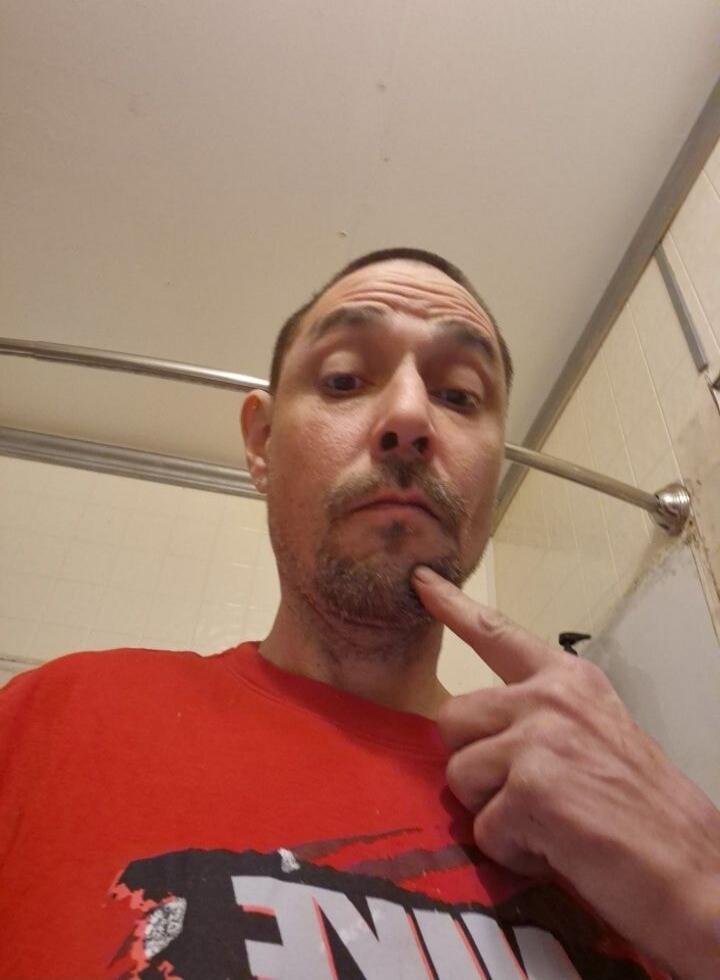 The image shows a man in a red t-shirt, with a beard and mustache, pointing to his chin. The background appears to be a bathroom with a shower curtain rod and tiled walls. There is no discernible speech in the image.