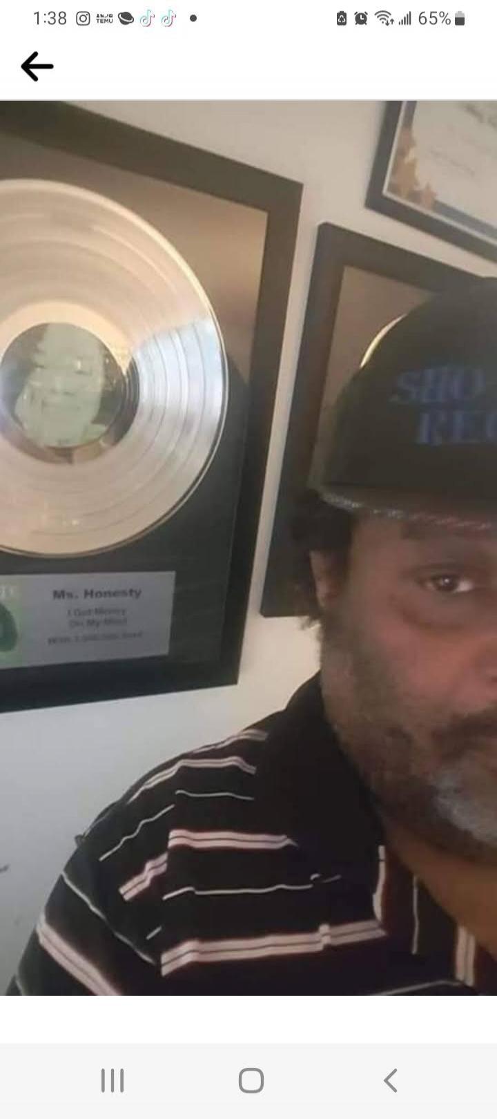 A man with a beard and a cap is in the foreground. In the background, a framed silver record award is visible. The award text reads: 'Ms. Honesty', 'I Got Money', 'On My Mind', 'With Lyrical Soul'. The man's cap has 'SHO-REC' written on it.