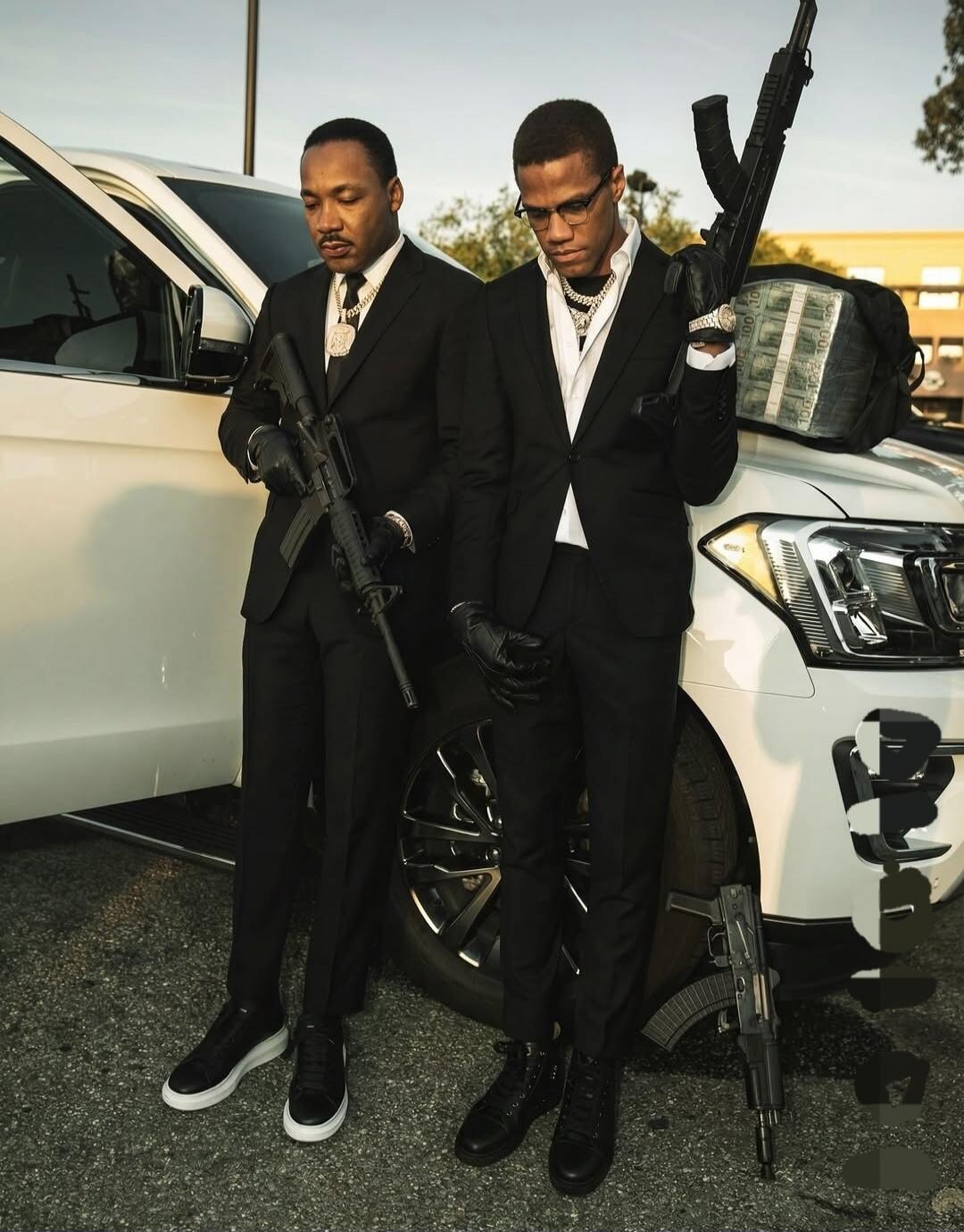 Two men in suits standing beside a white SUV, each holding rifles.