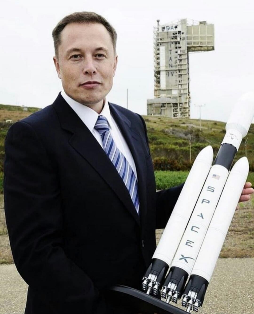 Elon Musk holding a SpaceX Falcon Heavy rocket model. In the background, a launch tower with 'USA' and 'TITAN IV' text is visible. The rocket model also has 'SPACEX' and a 'USA' flag.