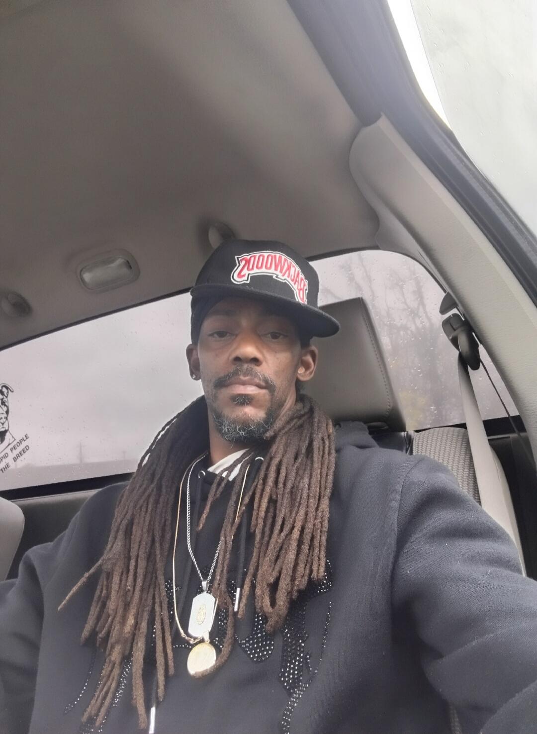 A man with dreadlocks and a beard is sitting in a car, taking a selfie. He is wearing a black hoodie and a black baseball cap with 'BACKWOODS' written in red. A sticker on the back window reads 'STUPID PEOPLE THE BREED' next to a dog image.