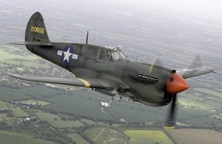 A vintage military aircraft, likely a P-40 Warhawk, flying in the sky over green fields. The aircraft is dark green with a red nose cone and a white star insignia on its wing. A pilot is visible in the cockpit. The tail number '210855' is visible.