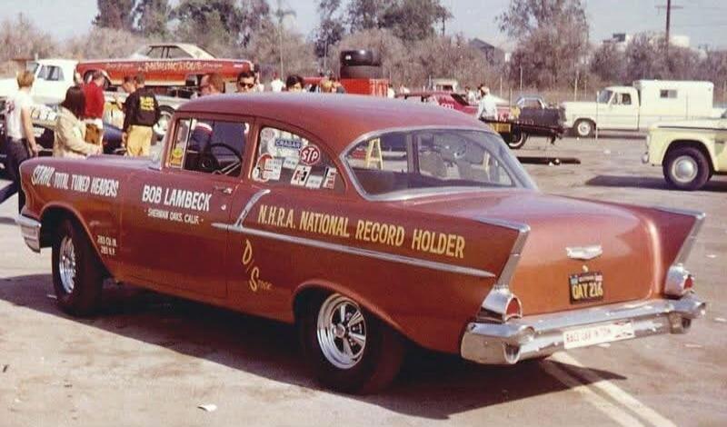 A vintage drag racing car, a red 1957 Chevrolet Bel Air, is parked at what appears to be a drag strip or car show. The car has various racing decals and text on its side, including 