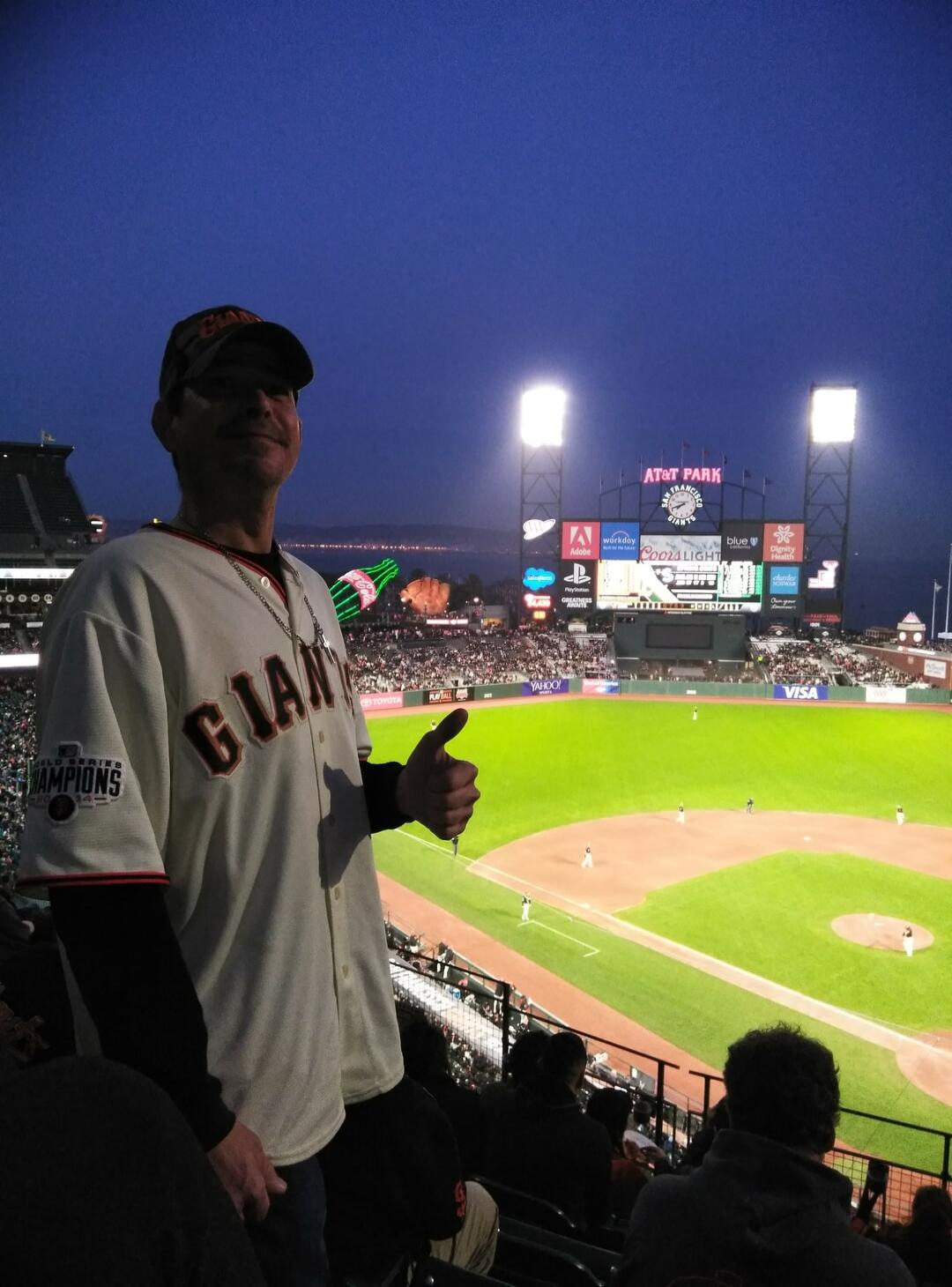 A man wearing a San Francisco Giants jersey and cap is giving a thumbs-up at a baseball game in AT&T Park. The jersey has a 'World Series Champions 2014' patch. The scoreboard shows various advertisements including Coors Light, Workday, Adobe, and PlayStation. A large neon sign of a Coca-Cola bottle and a baseball glove is visible in the background
