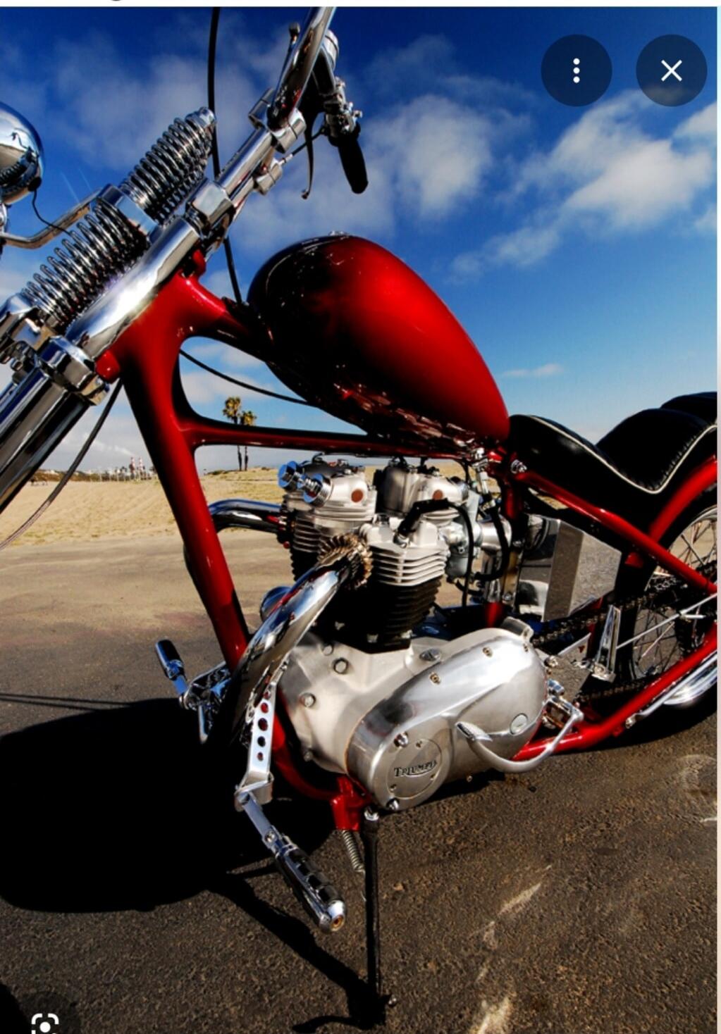 Close-up of a red motorcycle with a visible engine and fuel tank, parked on a paved surface with a beach and blue sky in the background.