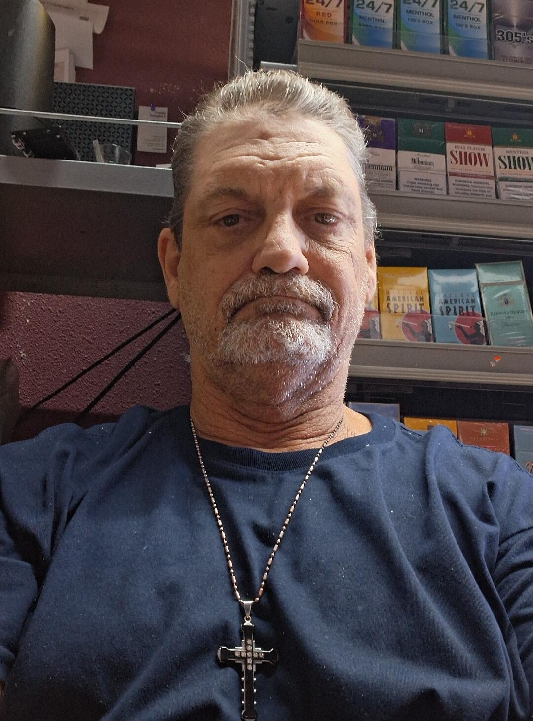 A man with gray hair and a beard, wearing a dark blue t-shirt and a cross necklace, takes a selfie in front of shelves stocked with various cigarette brands. Visible brands include 24/7, Menthol, Millennium, Show, American Spirit, and Benson & Hedges. A warning about cigarettes causing cancer and heart disease is also visible.