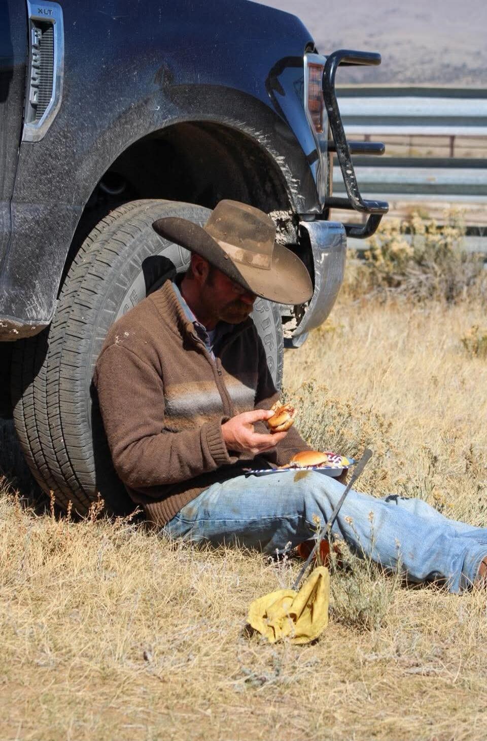 XLT. A man in a cowboy hat is sitting on dry grass, leaning against a pickup truck tire, eating a burger. Another burger is on a plate on his lap.