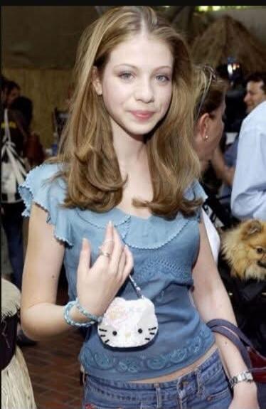 A young woman with long brown hair, wearing a blue top and jeans, holding a small Hello Kitty purse. She is smiling slightly. Other people and a small dog are visible in the background.