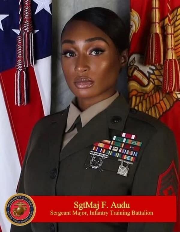 A woman in a military uniform, identified as SgtMaj F. Audu, Sergeant Major of the Infantry Training Battalion, stands in front of the US flag and a Marine Corps banner. Her uniform displays various ribbons and medals. The text on the banner reads: 'DEPARTMENT OF THE NAVY UNITED STATES MARINE CORPS SgtMaj F. Audu Sergeant Major, Infantry Training B