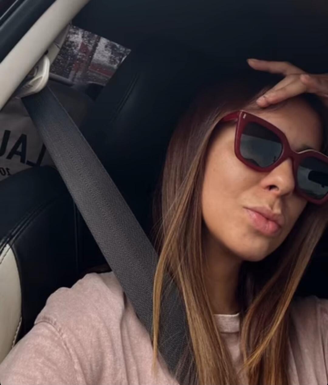 A woman in a car wearing sunglasses and a seatbelt, making a kissy face. Her hand is on her head.