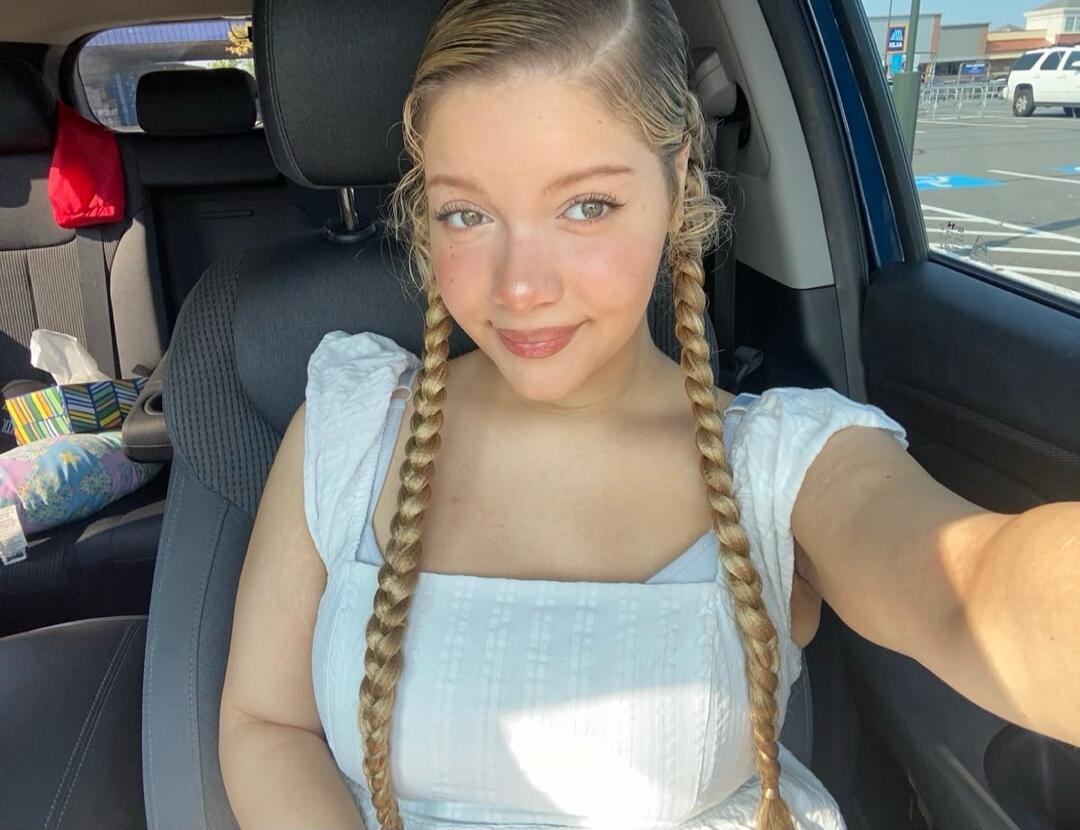 A young woman with blonde braids is sitting in a car, smiling. In the background, a parking lot with an Aldi store sign is visible.