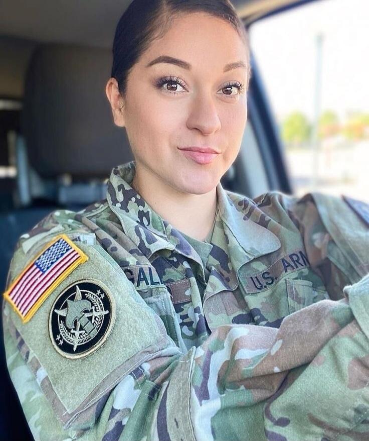 A woman in a US Army camouflage uniform is smiling while sitting in a car. Patches on her sleeve include the American flag and a military insignia. The text 'U.S. ARM' is visible on her uniform.