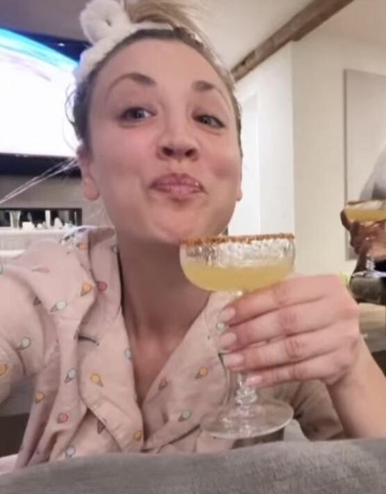 A woman in pajamas and a bunny ear headband holds a cocktail with a salted rim, making a playful face.