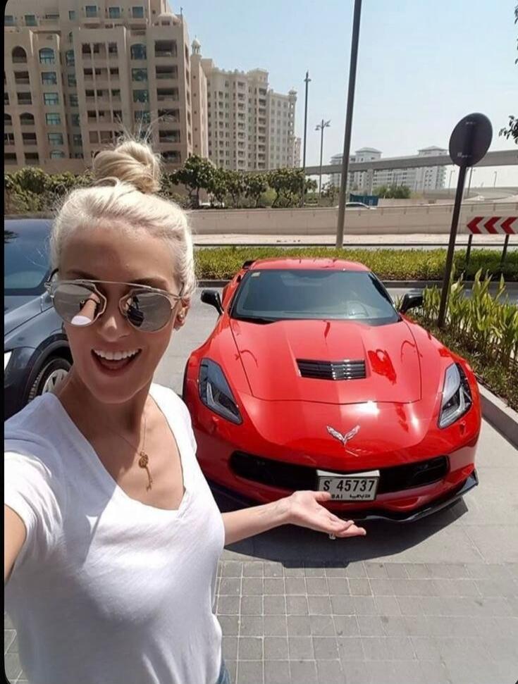 A blonde woman in sunglasses smiles and gestures towards a red sports car, possibly a Chevrolet Corvette, parked in an urban setting with modern buildings in the background. The car has a Dubai license plate with numbers, Latin letters 'BAI', and Arabic script.