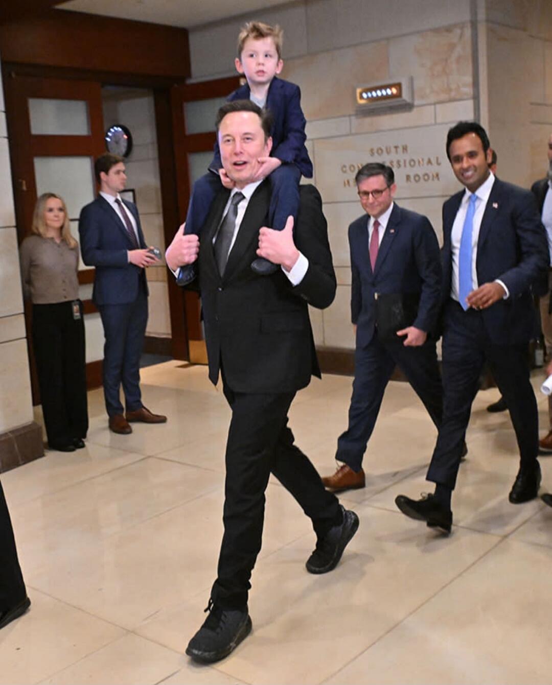 Elon Musk carrying his son on his shoulders in a building with a sign 'SOUTH CONGRESSIONAL ROOM'.