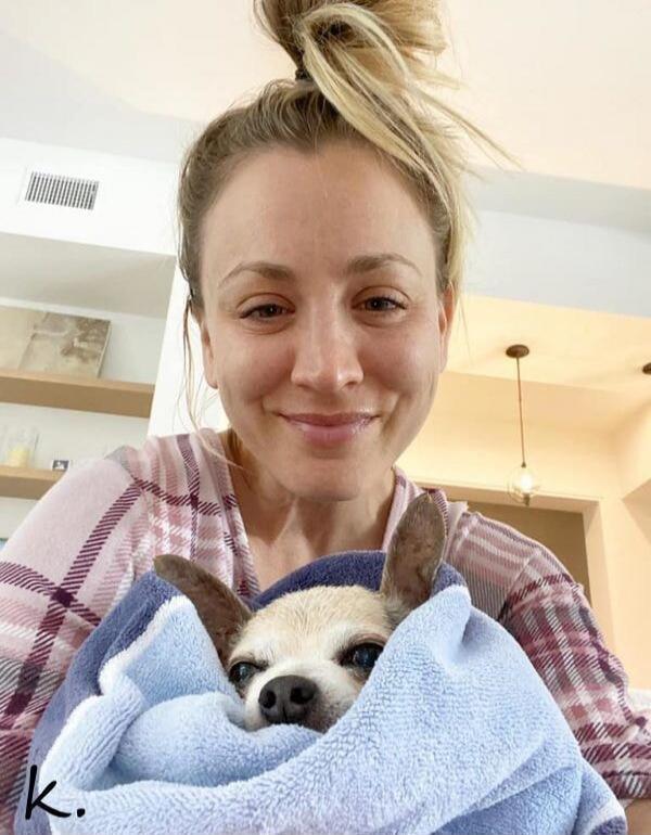 A woman with her hair in a messy bun, smiling at the camera, holds a small dog wrapped in a blue and white towel. The dog peeks out from the towel with a slightly grumpy expression. There is a small 'k.' in the bottom left corner.