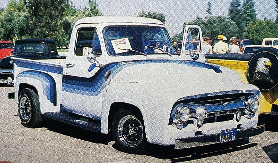 A white vintage Ford pickup truck with blue gradient stripes is parked outdoors. The license plate reads 'MIKE 54'. In the background, other vehicles and people are visible, including a yellow truck with a spare tire cover featuring an eagle design.