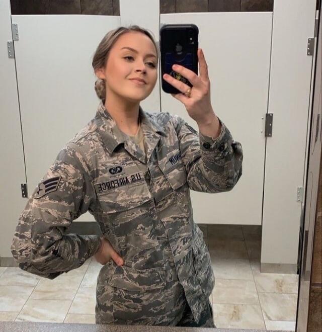 A young woman in a US Air Force uniform takes a selfie in a mirror. The text 'AIR FORCE' and 'NOW' are visible on her uniform, though mirrored.
