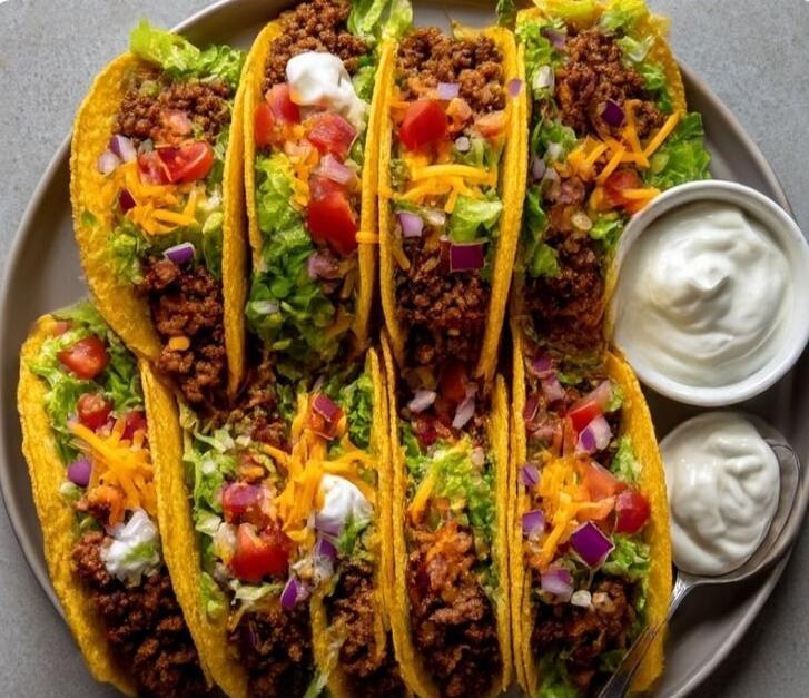 A plate of delicious hard-shell tacos filled with ground beef, shredded lettuce, diced tomatoes, red onions, shredded cheese, and a dollop of sour cream. Two small bowls of sour cream are visible on the side.