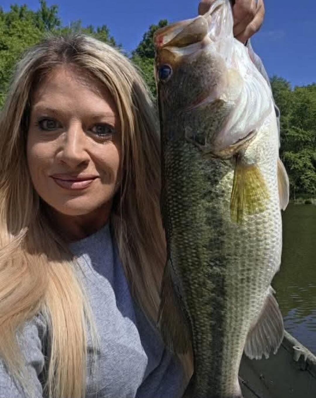 A woman with long blonde hair is holding a large fish, possibly a largemouth bass, in front of her. She is smiling slightly. The background shows a body of water, green trees, and a clear blue sky, indicating an outdoor fishing scene.