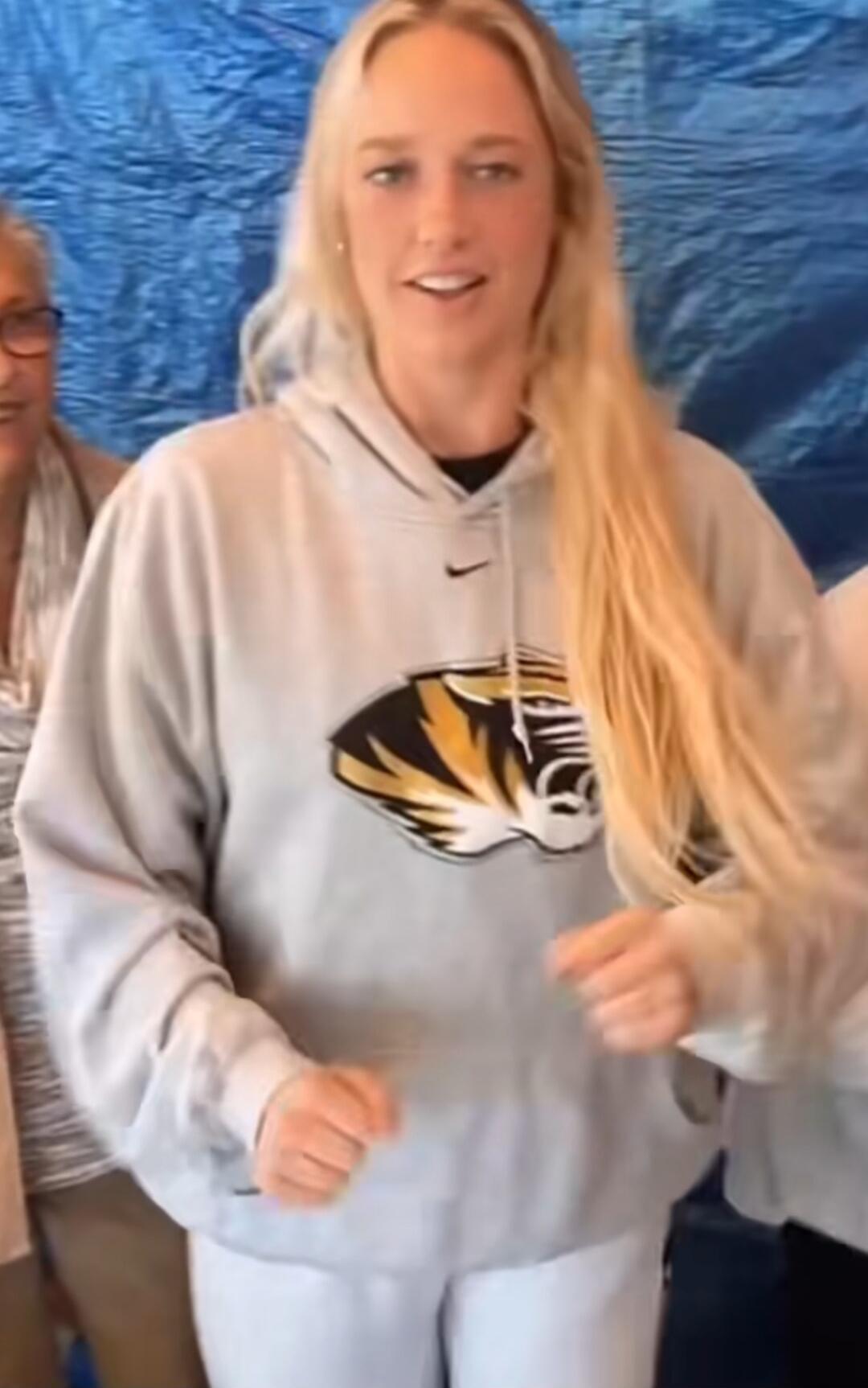 A young woman with long blonde hair is smiling and looking at the camera. She is wearing a grey hoodie with a black and gold tiger logo, likely representing the University of Missouri (Mizzou). Another person is partially visible on the left side of the frame. The background is a blue textured surface.