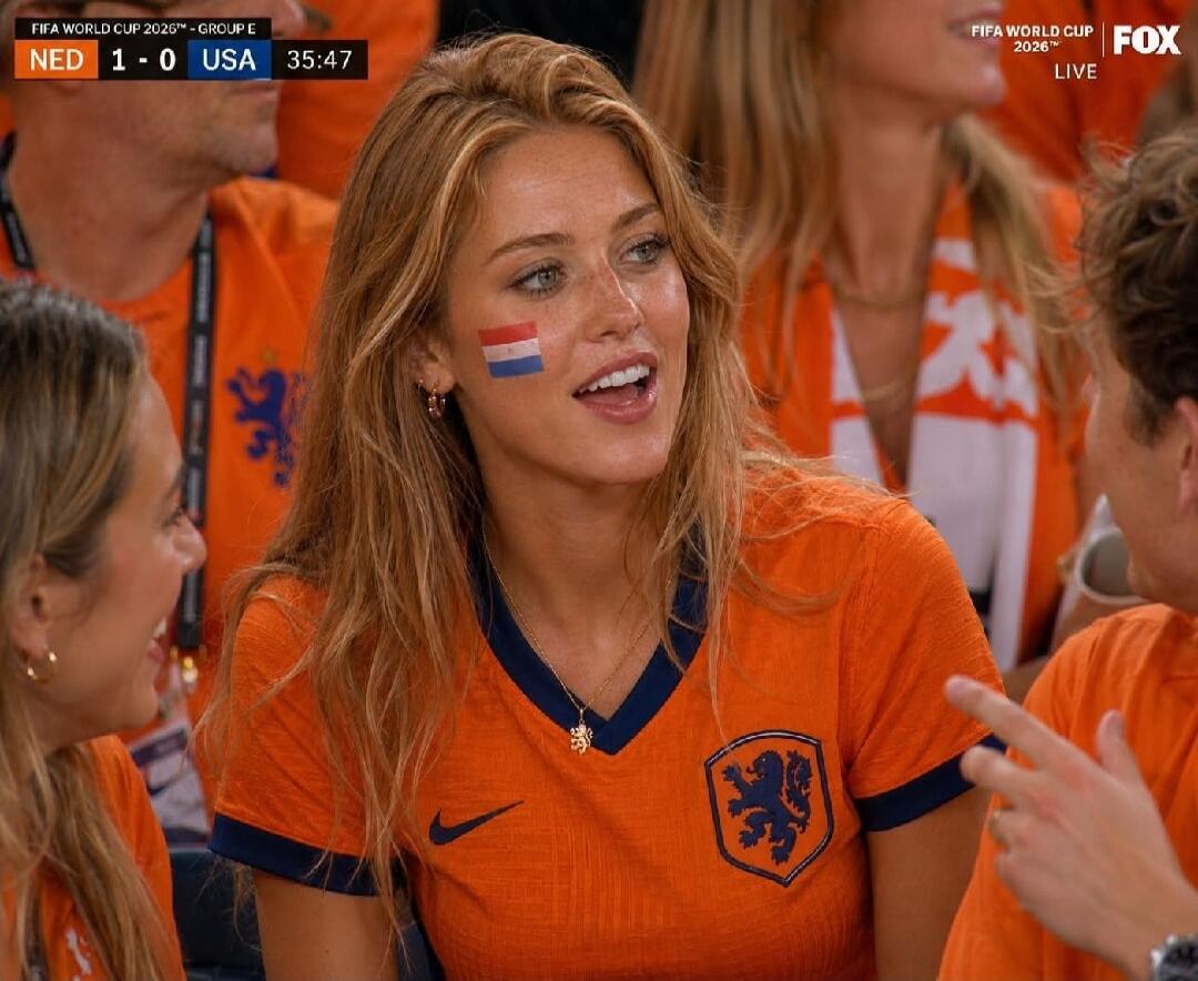 FIFA WORLD CUP 2026™ - GROUP E, NED 1 - 0 USA, 35:47, FIFA WORLD CUP 2026™, FOX, LIVE. A young woman with long blonde hair, wearing an orange Netherlands football jersey, with a Dutch flag painted on her cheek, smiling and watching a football match.